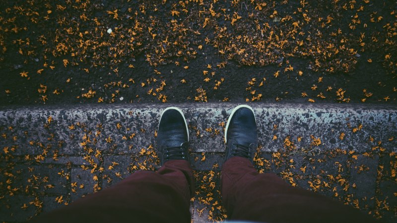 A person's dark shoes beside a kerb scattered with fallen leaves