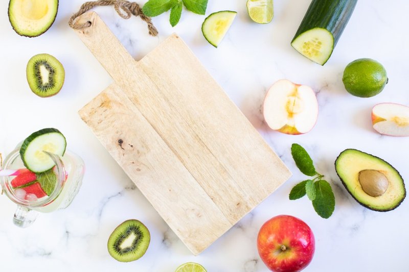 Avocado, cucumber and other fresh produce around a wooden chopping board