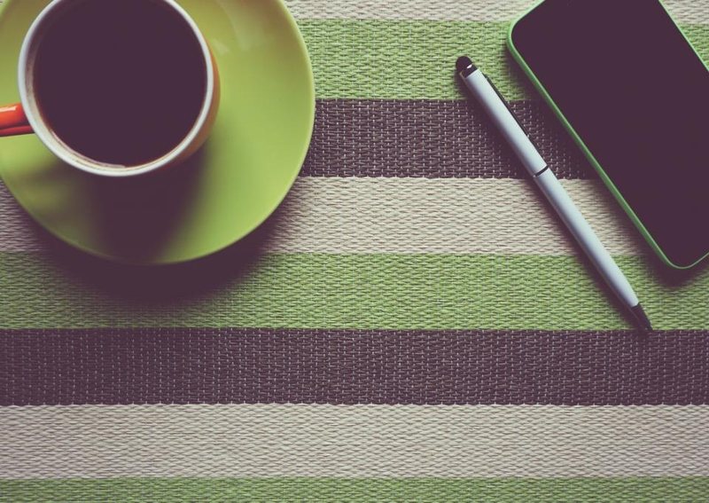 A green cup of black coffee beside a phone and pen on a striped placemat