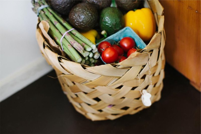 A woven shopping basket holding asparagus, tomatoes, peppers and avocados