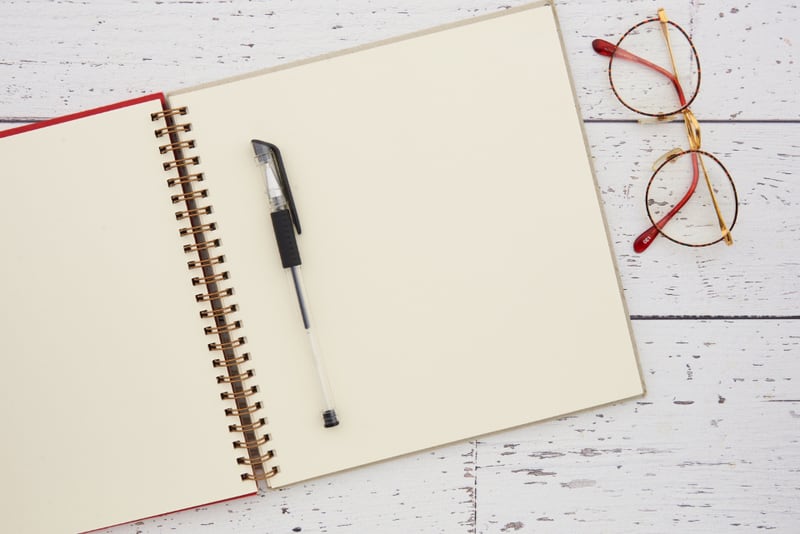 A blank spiral notebook with a pen and reading glasses on a pale wooden surface
