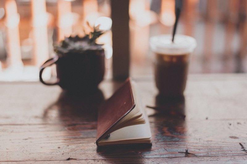 An open notebook beside a small potted plant and a drink on a wooden table