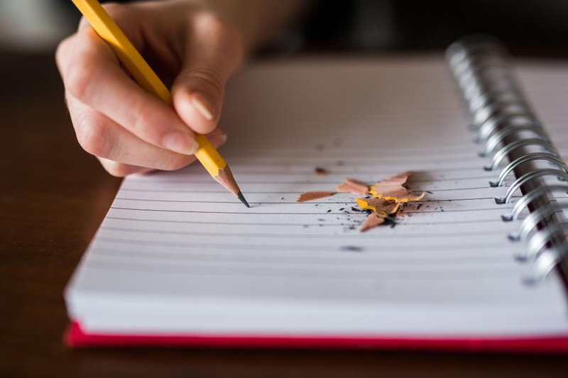 A hand holding a pencil over a blank notebook beside pencil shavings